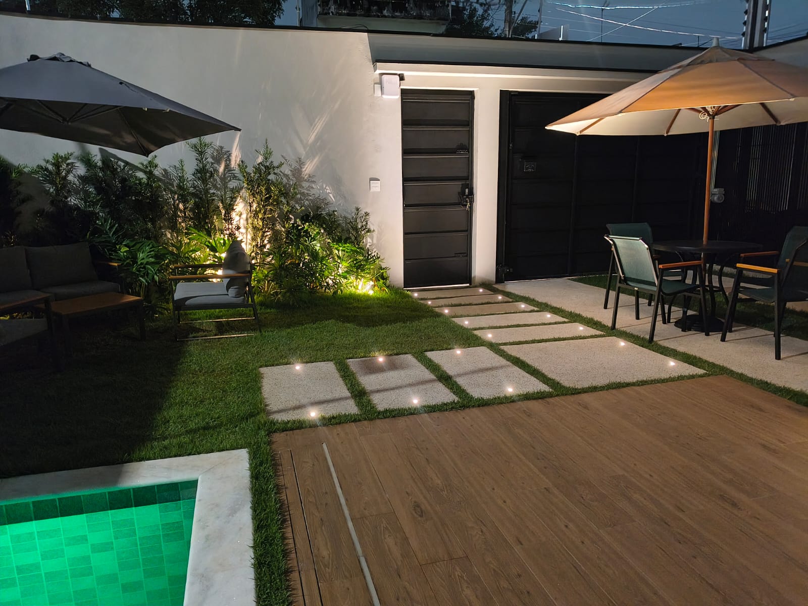 Área externa iluminada com deck, jardim e piscina à noite
